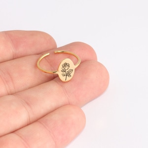Può includere: Anello regolabile color oro con un elemento centrale ovale inciso con una rosa dettagliata. La fascia aperta consente la regolazione della taglia. Il disegno della rosa è nero.