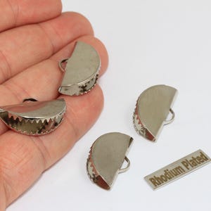 Puede incluir: Cuatro terminales de metal plateado para cintas con forma semicircular y dientes serrados para sujetar la tela. Cada pieza cuenta con una pequeña anilla en la parte superior para su fijación. Se incluye una etiqueta con el texto Rhodium Plated.