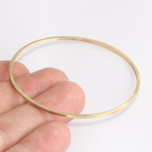 May include: A thin, gold-colored bangle bracelet. The circular bracelet is made of a thin metal band and is displayed against a white background. The bracelet is held by a hand.