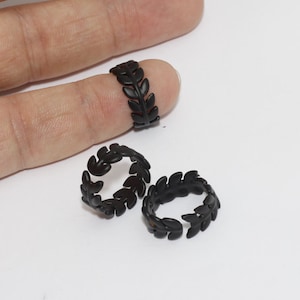 Puede incluir: Tres anillos negros con forma de hojas. Un anillo está en un dedo y dos están sobre una superficie blanca. Los anillos tienen un diseño delicado inspirado en la naturaleza, con hojas estilizadas.