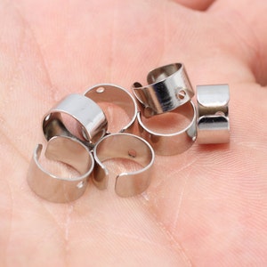Könnte beinhalten: Eine Sammlung von silberfarbenen, offenen Ohrbügelrohlingen aus Metall. Jede Manschette ist ein kleines, gebogenes Band mit einem kleinen Loch. Die Manschetten werden auf einem hellfarbigen Hintergrund angezeigt.