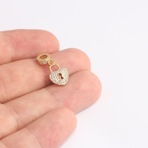 May include: A gold-coloured heart-shaped charm with a keyhole design, encrusted with small, clear crystals. The charm is suspended from a small, round, crystal-studded loop, and is likely a jewellery component.