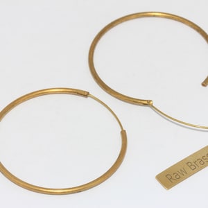 May include: Two gold-colored hoop earrings with a simple design. One earring is open, showing the clasp. A small, rectangular tag with the words "Raw Brass" is also present. The earrings are on a white background.