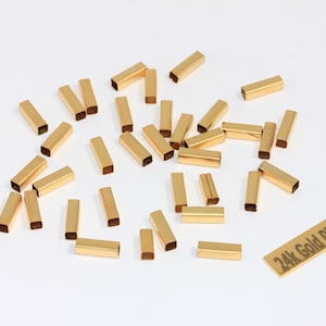 Puede incluir: Un conjunto de pequeños tubos rectangulares de metal chapado en oro dispersos sobre una superficie blanca. Los tubos están abiertos por ambos extremos. También hay un pequeño letrero rectangular con el texto "24k Gold Plated".