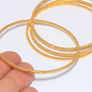 Puede incluir: Un conjunto de brazaletes dorados con textura martillada. Los brazaletes circulares son de varios tamaños, uno de ellos sostenido por una mano. Las joyas se exhiben sobre un fondo blanco.