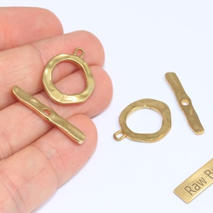 May include: A collection of raw brass jewelry findings, including two toggle clasps, two rings, and a rectangular tag with the text "Raw Brass". The pieces have a hammered texture and a warm, golden color.