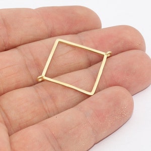 May include: A gold-colored, open-frame diamond-shaped charm with small loops at two corners. The charm is held in a hand against a white background. This jewelry component is suitable for crafting earrings, necklaces, or other accessories.