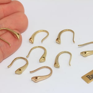 May include: A selection of gold-coloured raw brass earring hooks. The hooks have a curved design with a small hole at the end. A small rectangular tag with the text "Raw Brass" is also present.