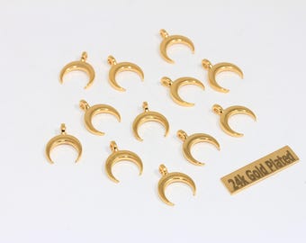 May include: A collection of gold-colored crescent moon charms, each with a small loop at the top for stringing. A small rectangular tag reads "24k Gold Plated". The charms are scattered on a white surface.