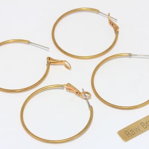 May include: Four gold-colored hoop earrings with a simple design are displayed on a white surface. Each earring has a hinged closure. A small, rectangular tag with the words "Raw Brass" is also present. The earrings are a jewelry item.