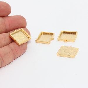 Puede incluir: Bandejas cuadradas doradas para colgantes con superficies texturizadas. Las bandejas tienen un pequeño lazo para sujetarlas a una cadena u otro componente de joyería. La imagen muestra las bandejas sobre un fondo blanco.