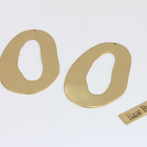 May include: Two gold-coloured, abstract-shaped brass earring components with a hole for hanging. A small rectangular brass tag with the text "Raw Brass" is also present. The earrings have an organic, open design.