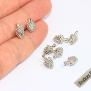 Könnte beinhalten: Silberfarbene, rhodiniertes Kiefernzapfen-Charms. Diese kleinen, detaillierten Anhänger haben eine strukturierte Oberfläche und eine Schlaufe zum Befestigen an Schmuck. Ein kleines Metallschild mit dem Text "Rhodium Plated" ist ebenfalls sichtbar.