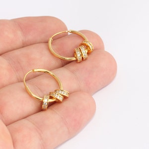May include: A pair of gold-colored hoop earrings. Each earring features a small hoop with three decorative, sparkling bands of clear crystals. The earrings are displayed against a neutral background.