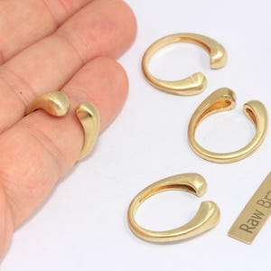 May include: A collection of gold-colored brass rings with a unique open design. One ring is worn on a finger, while three others are displayed nearby. A small rectangular tag with the text "Raw Brass" is also present.
