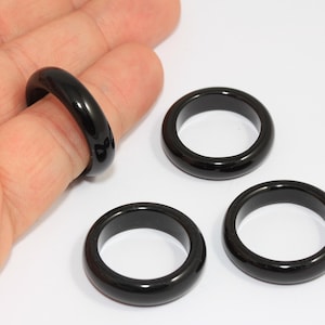 May include: A collection of black rings, one worn on a finger, with a glossy finish. The rings are round and appear to be made of a solid, dark material. The background is plain white, highlighting the jewelry.