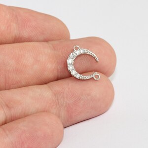 May include: A silver-toned crescent moon charm, encrusted with clear, sparkling crystals. The charm has two small loops for attaching to jewelry and is held in a person's hand against a white background. This charm is suitable for jewelry making.