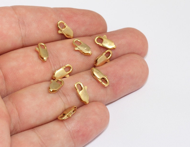 May include: A collection of small, gold-colored lobster claw clasps. These jewelry findings are designed with a curved body and a spring-loaded clasp mechanism. The clasps are scattered against a white background.