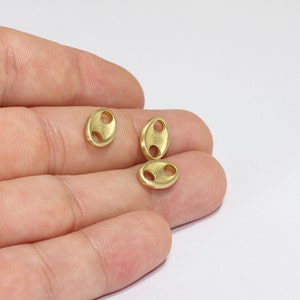 May include: Four small, gold-colored, oval-shaped metal beads. Each bead has two circular holes. The beads are scattered on a light background. These beads are likely used for jewelry making or other craft projects.