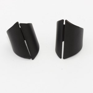 May include: Two matte black cuff bracelets are displayed on a white background. Each bracelet has a curved, open design with a vertical slit. The bracelets are made of a solid material and have a smooth finish.