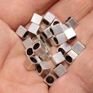 May include: A selection of small, silver square beads, each with a central hole. The beads are held in a hand, and the metallic surfaces reflect light. These beads are likely for jewellery making or other crafts, and are measured in centimetres.