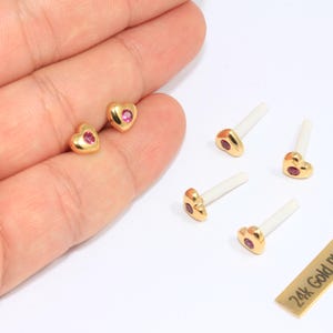 May include: Gold-plated heart-shaped stud earrings with a small pink gemstone in the centre. The earrings are displayed with several earring posts. A small gold rectangle reads "24k Gold Plated".