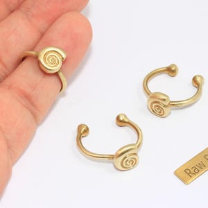 Puede incluir: Anillos y puños de oreja de latón dorado con un diseño en espiral. Un anillo está en un dedo y se ven dos puños de oreja. También hay una pequeña etiqueta de latón con el texto "Raw Brass".