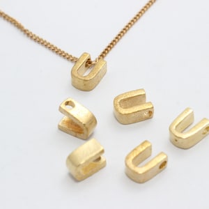May include: A gold-colored necklace with a 'U' pendant, along with several loose 'U' shaped charms. The charms and necklace chain are a warm, metallic tone, set against a white background. The pendant and charms have a matte finish.