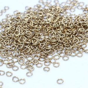 May include: A collection of small, gold-coloured jump rings, scattered on a white surface. The rings are circular and appear to be made of metal. They are likely used for jewellery making or other crafts.