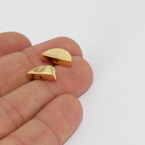 May include: Two gold-colored, half-moon shaped metal beads. Each bead has a flat, rectangular base and a curved top with a small hole. The beads are displayed against a white background.