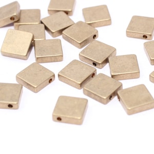 May include: A collection of small, square, brass-colored beads scattered on a white surface. Each bead has a flat, square shape with a small hole drilled through the center. The beads are evenly colored and appear to be made of metal.
