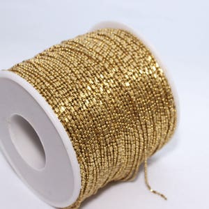 May include: A spool of gold-colored beaded chain. The chain is tightly wound around a white plastic spool. The beads are small and rectangular, creating a textured surface. The background is plain white.