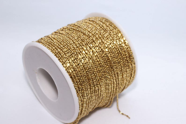 May include: A spool of gold-colored beaded chain. The chain is tightly wound around a white plastic spool. The beads are small and rectangular, creating a textured surface. The background is plain white.