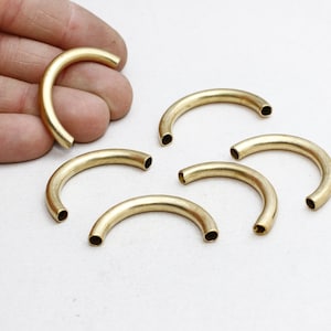 May include: Several curved, brass-colored metal tubes are displayed on a white surface. One tube is held by a hand. The tubes have a hollow, cylindrical design, and are likely jewelry components or craft supplies.