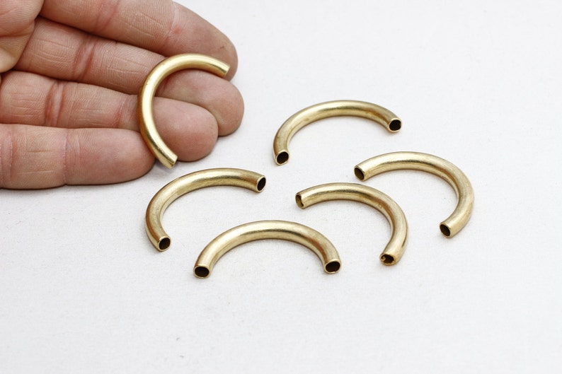 May include: Several curved, brass-colored metal tubes are displayed on a white surface. One tube is held by a hand. The tubes have a hollow, cylindrical design, and are likely jewelry components or craft supplies.