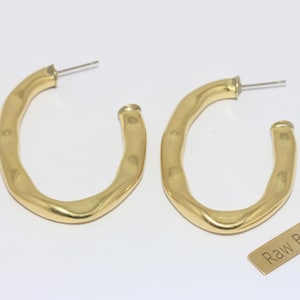 May include: A pair of gold-colored, hammered brass hoop earrings. The earrings have a unique, irregular shape and are displayed on a white background. A small tag that reads "Raw Brass" is also present.