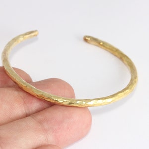Op de afbeelding: Een goudkleurige manchetarmband met een gehamerde textuur. De open armband heeft een eenvoudig, elegant ontwerp. De armband wordt door een hand tegen een witte achtergrond gehouden. De manchet heeft een diameter van ongeveer 6,35 cm.