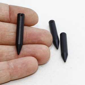 May include: Three black, cylindrical pendants with a pointed end and a small hole at the top. One pendant is held in a hand, while the other two are on a white surface. The pendants are made of a matte black material.