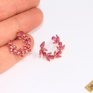 May include: A pair of gold-plated earrings with a floral design. Each earring features a half-circle arrangement of pink, teardrop-shaped gemstones. The earrings are displayed on a white surface.