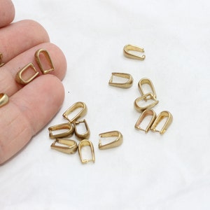 May include: A collection of small, gold-colored metal findings, shaped like a U with a loop at the top. These findings are scattered on a white surface, suggesting they are jewelry-making components or similar craft supplies.