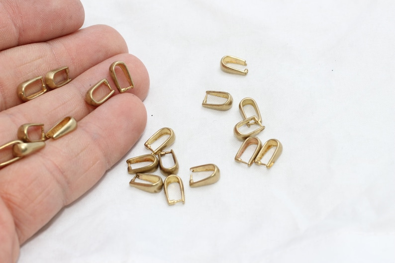 May include: A collection of small, gold-colored metal findings, shaped like a U with a loop at the top. These findings are scattered on a white surface, suggesting they are jewelry-making components or similar craft supplies.