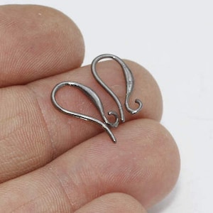 May include: Two silver-coloured earring hooks. The hooks are curved with a loop at the top and a straight wire at the bottom. The hooks are held in a person's hand against a white background. Jewellery making supplies.