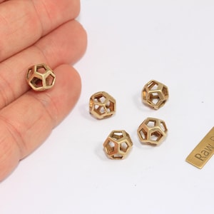 May include: A collection of small, geometric brass beads with an open, cage-like design. One bead is held in a hand, with four others and a small brass tag that reads "Raw Brass" on a white surface. These beads are likely for jewelry making.