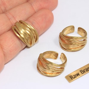 May include: Three gold-colored brass rings with a layered design. One ring is on a finger, and two are displayed separately. A small brass tag with the text "Raw Brass" is also present. The rings have a textured, organic look.
