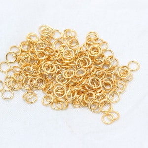 May include: A collection of small, gold-coloured jump rings, scattered on a white surface. These open rings are used for jewellery making and other crafts. The rings are round and have a shiny finish.