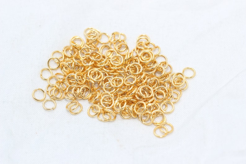 May include: A collection of small, gold-coloured jump rings, scattered on a white surface. These open rings are used for jewellery making and other crafts. The rings are round and have a shiny finish.