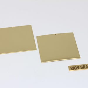 Puede incluir: Dos plantillas cuadradas de metal dorado con un pequeño agujero en la parte superior y una etiqueta rectangular que dice "RAW BRASS". Los artículos se muestran sobre una superficie blanca.