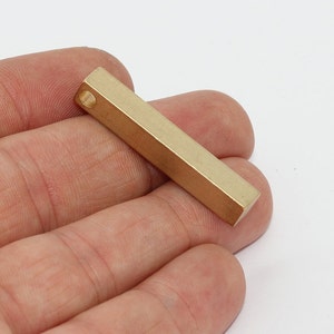 May include: A small, rectangular, gold-colored metal bar with a hole drilled at one end. The bar has a faceted design and is held in a person's hand. The object appears to be a jewelry component or a small decorative element.
