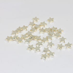 K&ouml;nnte beinhalten: Eine Ansammlung kleiner, metallisch goldfarbener sternf&ouml;rmiger Perlen, die auf einer schlichten Oberfl&auml;che verstreut sind. Jede Perle hat ein f&uuml;nfzackiges Sterndesign mit einer glatten, reflektierenden Oberfl&auml;che und einem kleinen Loch.