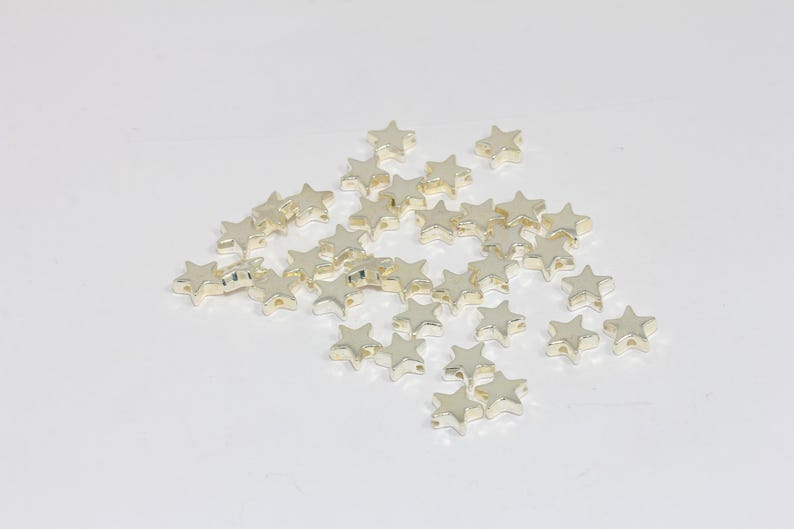 K&ouml;nnte beinhalten: Eine Ansammlung kleiner, metallisch goldfarbener sternf&ouml;rmiger Perlen, die auf einer schlichten Oberfl&auml;che verstreut sind. Jede Perle hat ein f&uuml;nfzackiges Sterndesign mit einer glatten, reflektierenden Oberfl&auml;che und einem kleinen Loch.